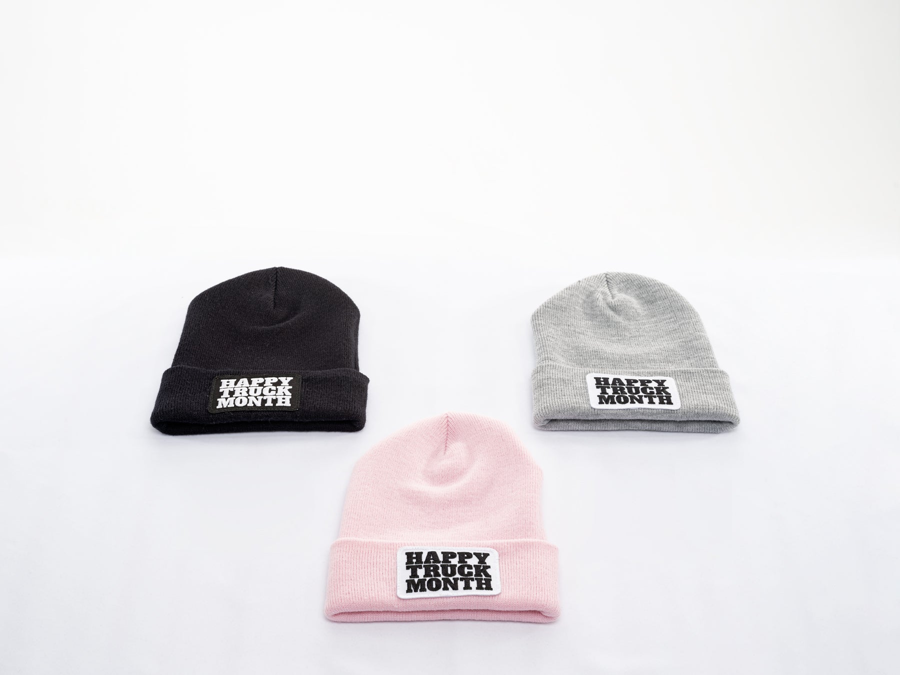 Three beanies in black, gray, and pink are arranged on a white surface. Each beanie has a patch on the front that reads HAPPY TRUCK MONTH in bold white and black letters.