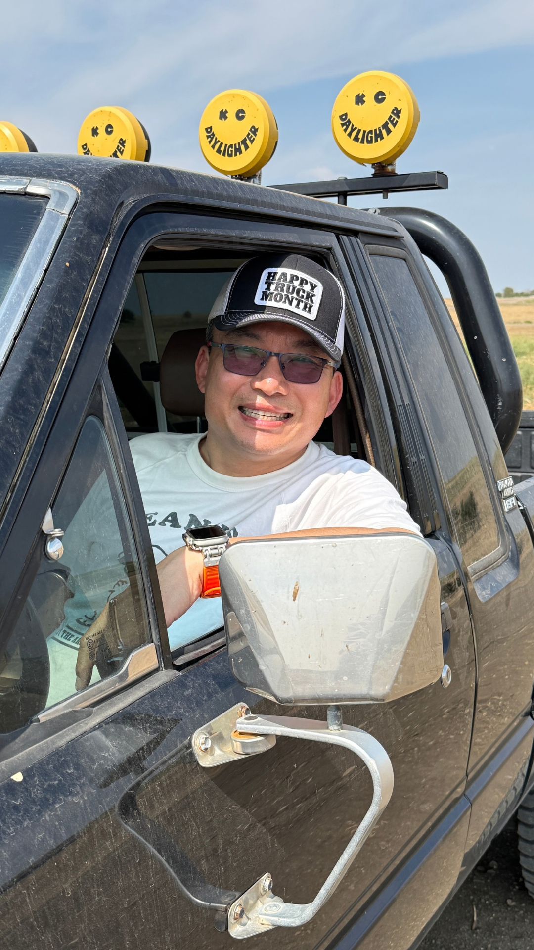 A man wearing a black and white 'Happy Hour is Night' baseball cap and sunglasses sits in the driver's seat of a black pickup truck, with yellow K&G Lighter roof lights visible above.