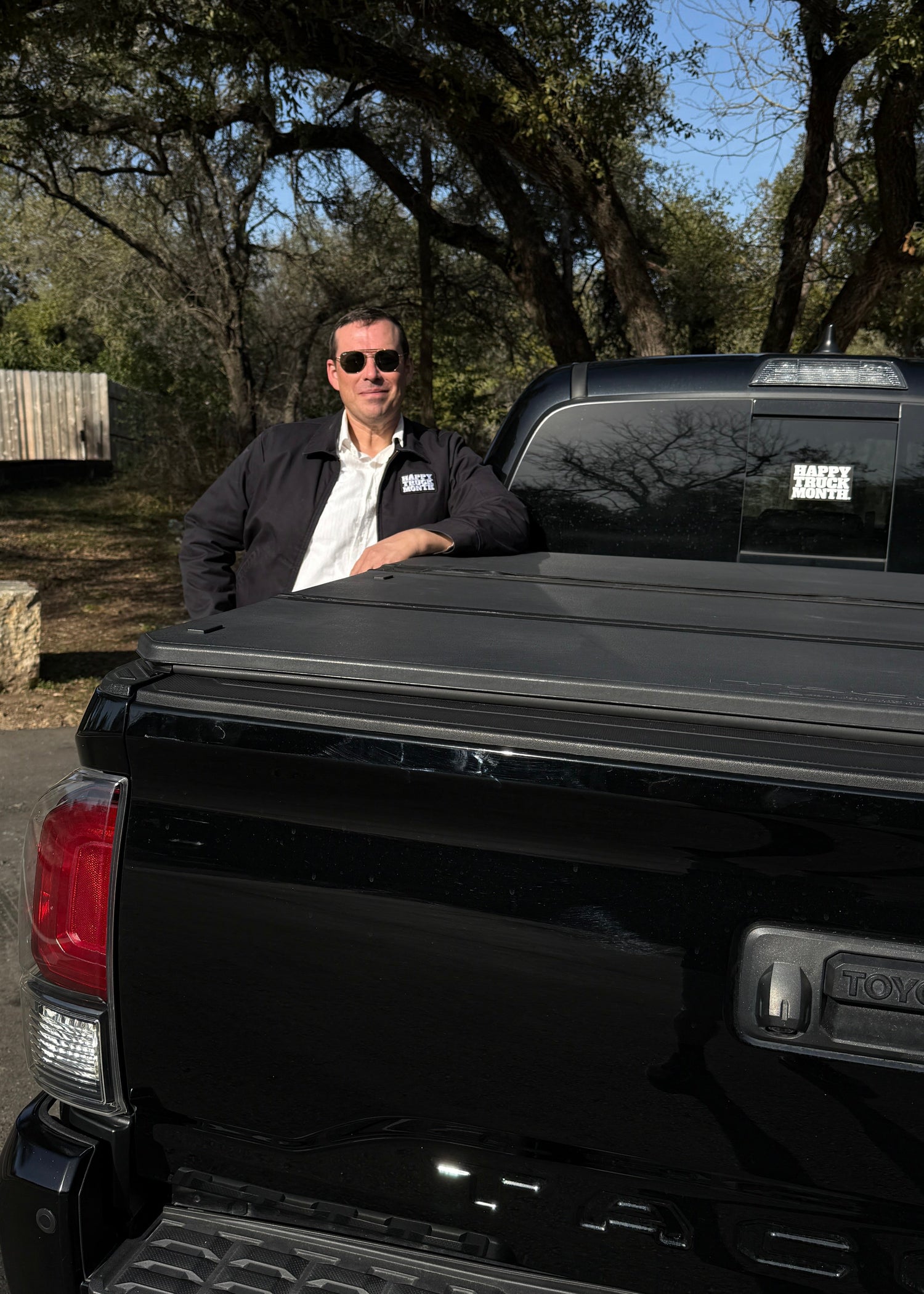 Man standing next to a black pickup truck with a tonneau cover in a wooded area.