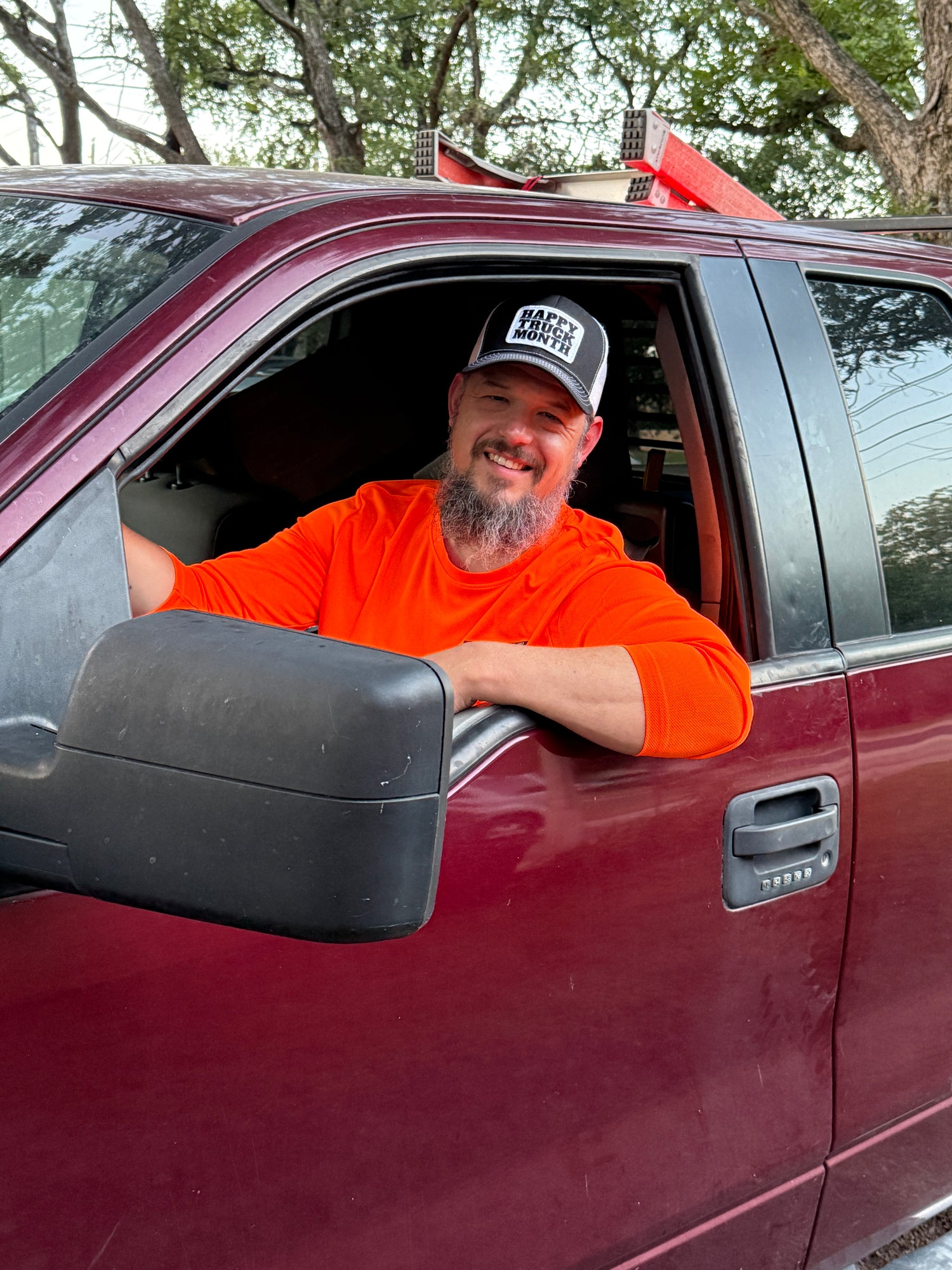 A smiling man with a beard wearing an orange t-shirt and a black and white 'RACE WARRIOR' cap leans out of the driver's side window of a maroon pickup truck, with trees in the background.