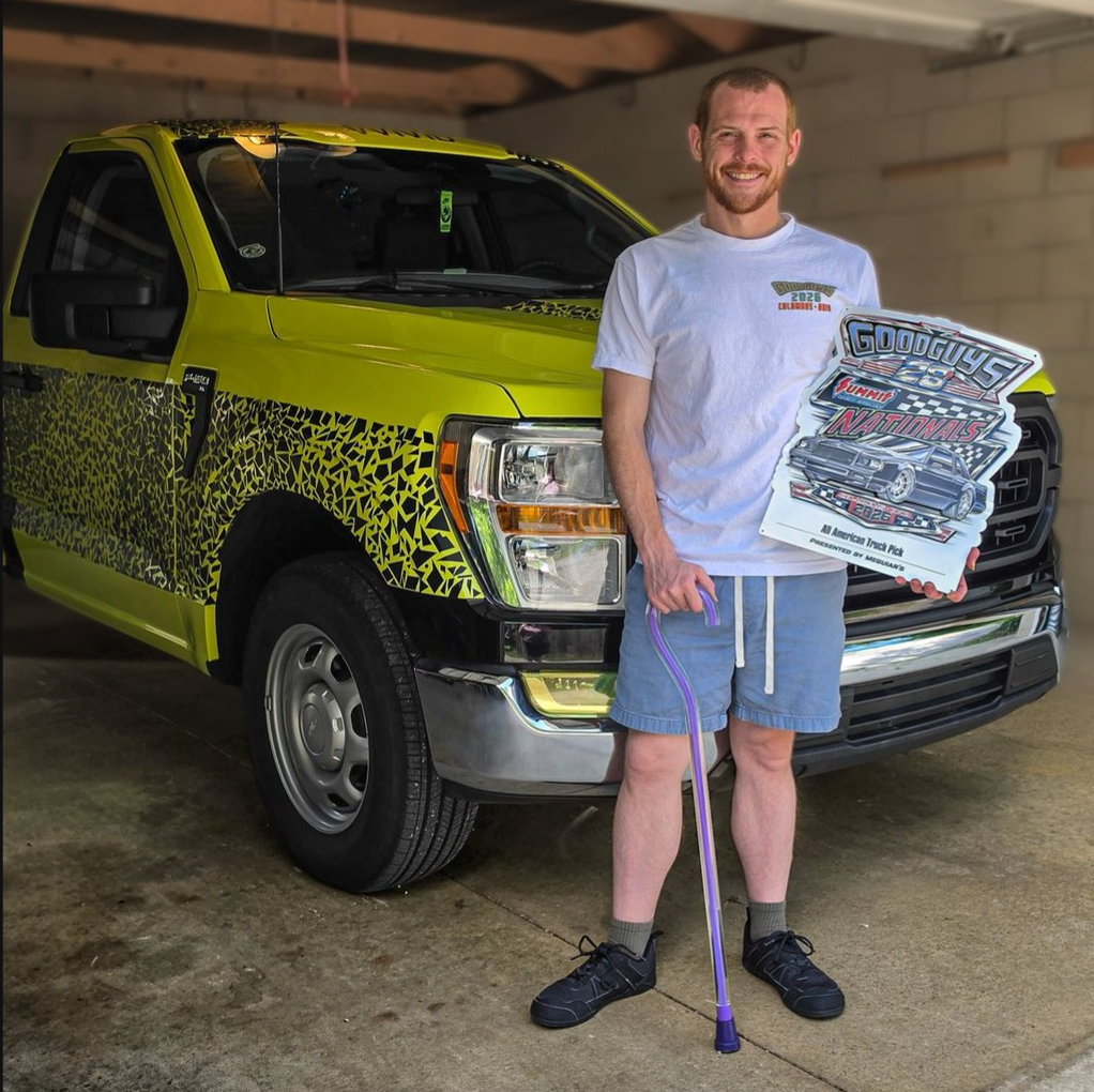 A man stands in a garage in front of a bright lime green pickup truck, holding a purple cane and a large printed sign that reads 'Good Luck with This Truck' and '4 Star Driver.' The truck has a bold black and yellow animal-print style wrap on the lower side panels.
