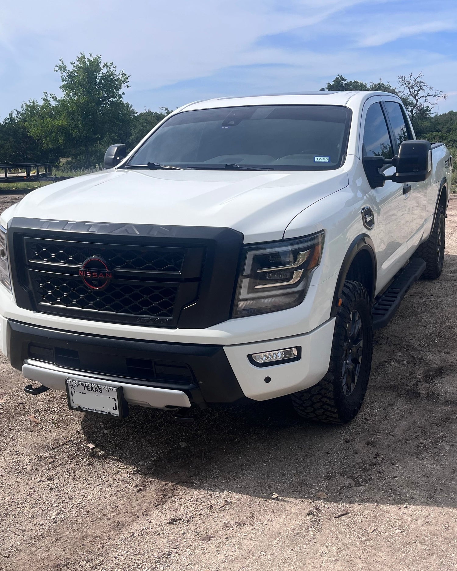 White Nissan Titan pickup truck 3/4 front view on gravel with trees and sky in background.