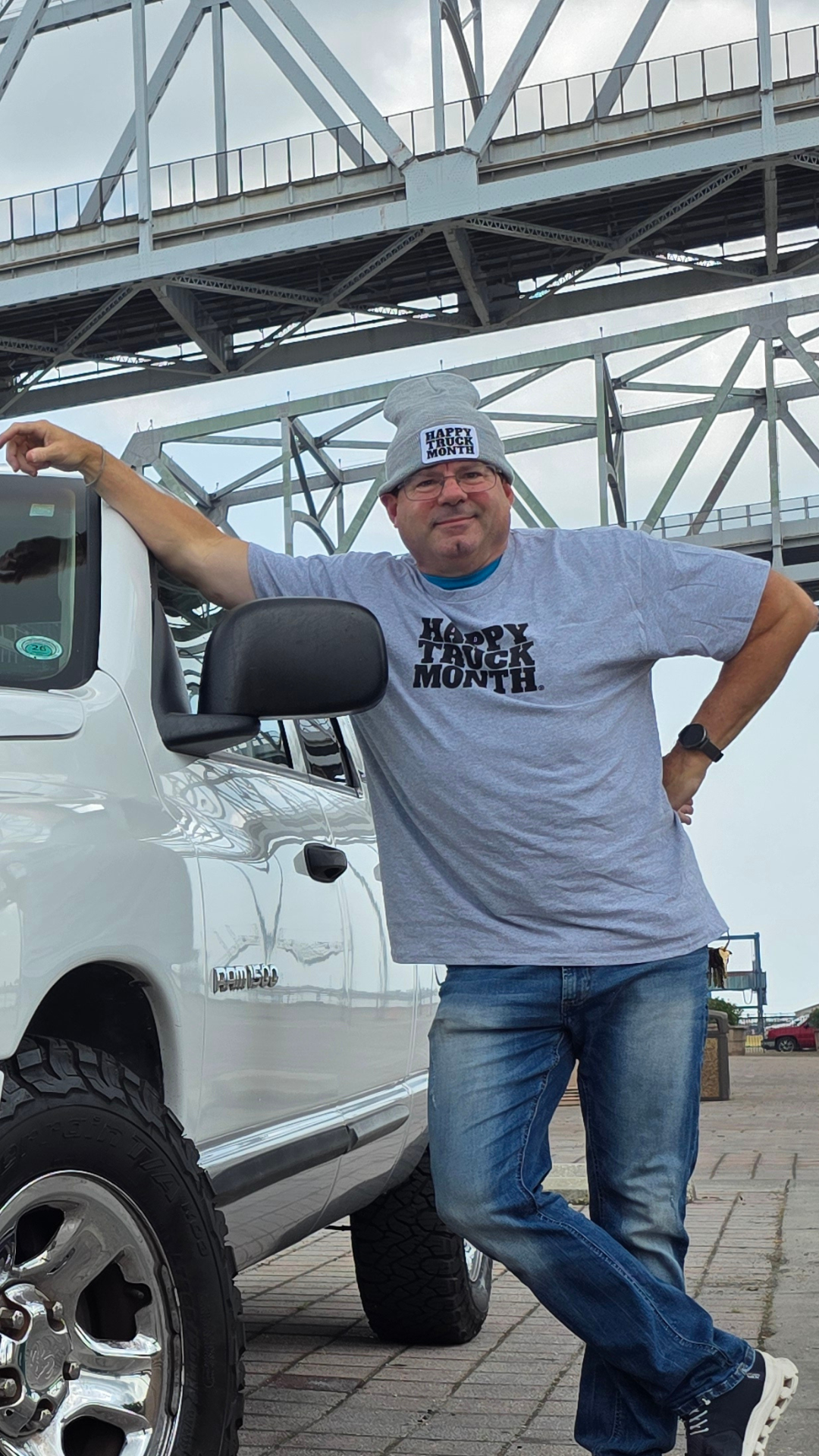 Man wearing a gray beanie and t-shirt with Happy Truck Month text, standing next to a white truck under a bridge.