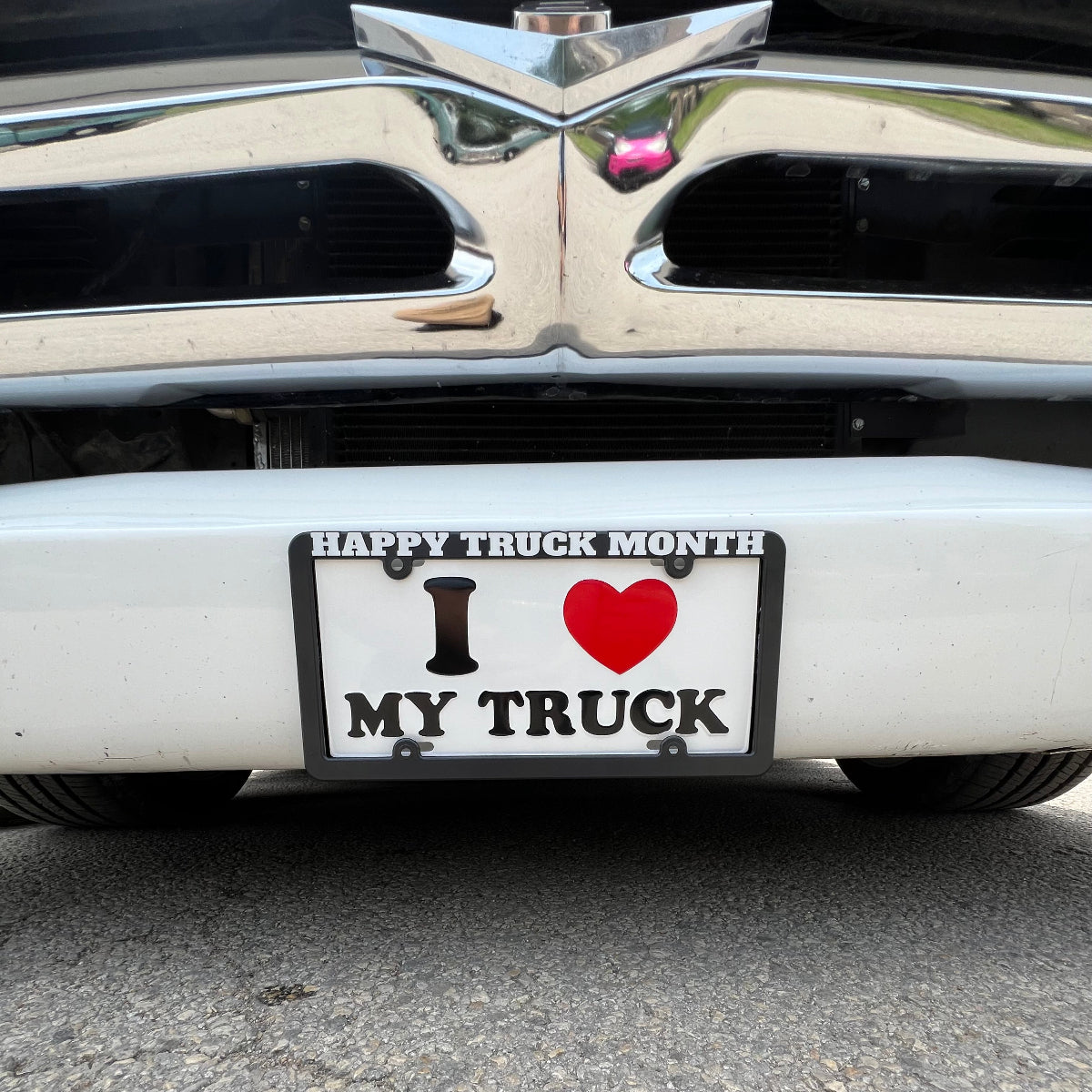 A product license plate frame reading "Happy Truck Month" in all caps framing a  novelty license plate that reads "I 'heart' my truck" in all caps on a classic white truck's front end. 