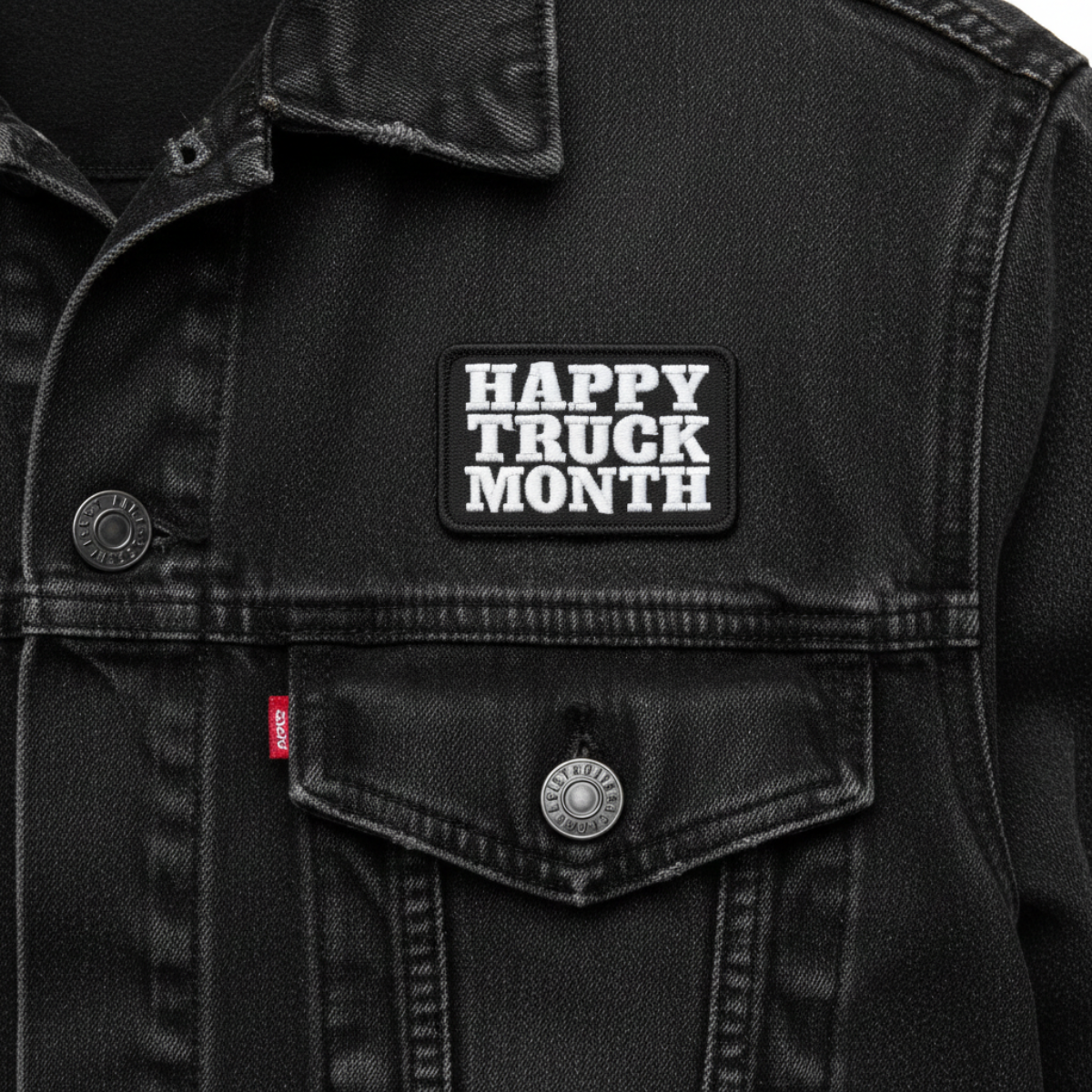 Close-up of a black denim trucker jacket with a rectangular black patch on the chest that reads 'HAPPY TRUCK MONTH' in white block letters. A small red tag is visible near the pocket, and the jacket has a dark washed denim texture.