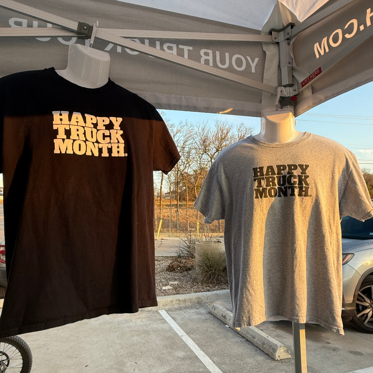 Two graphic t-shirts displayed on mannequins under a pop-up canopy at an outdoor market. One shirt is black with white text reading 'HAPPY TRUCK MONTH. The other shirt is gray with 'HAPPY TRUCK MONTH' in black text.