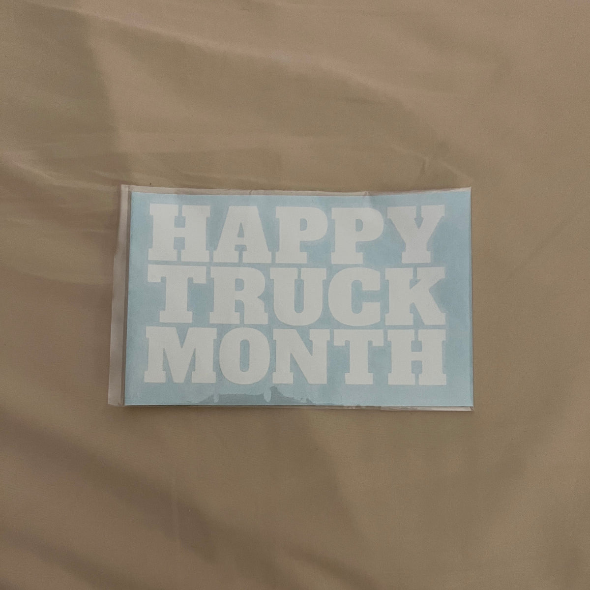 A rectangular light blue sticker or decal with the words 'HAPPY TRUCK MONTH' in large white block letters, placed on a beige fabric background.