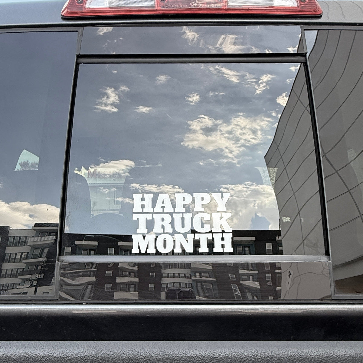 A white vinyl decal reading 'HAPPY TRUCK MONTH' applied to the rear window of a dark gray truck, with reflections of the sky and surrounding buildings visible on the glass.