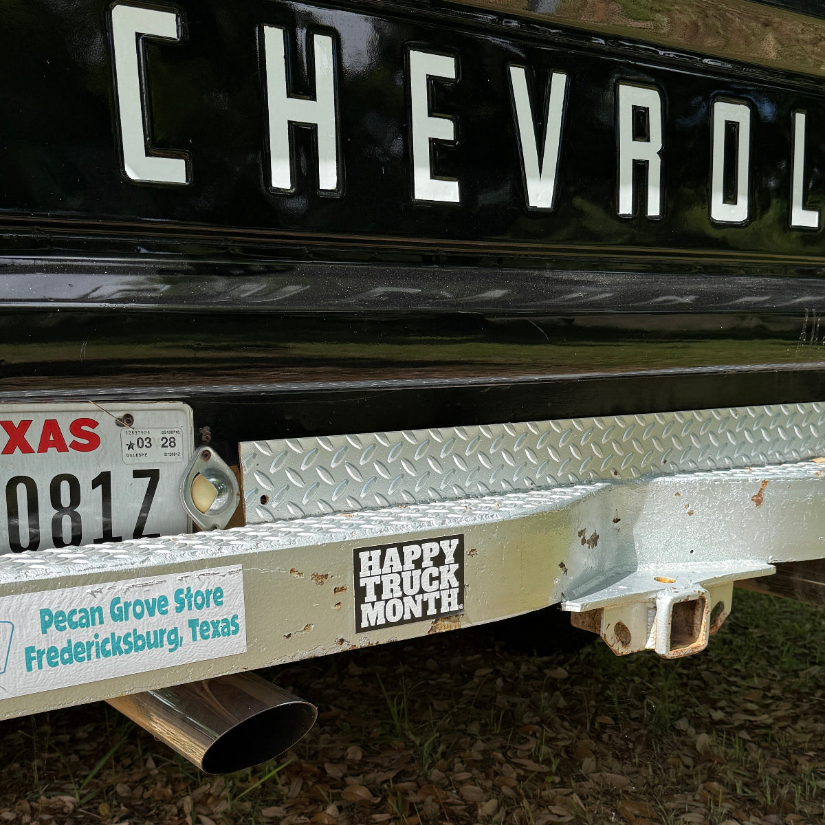 Close-up of the rear bumper and hitch area of a black Chevrolet truck outdoors on a leaf-covered ground. A Texas license plate, a 'Happy Truck Monthly' sticker, and part of the word 'CHEVROLET' are visible.