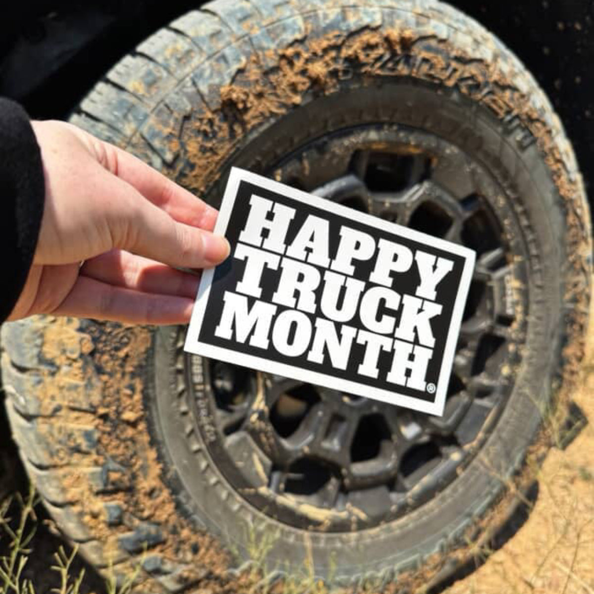 A hand holds a white rectangular sticker with bold black text reading 'HAPPY TRUCK MONTH.' in front of a muddy off-road tire on sandy ground.